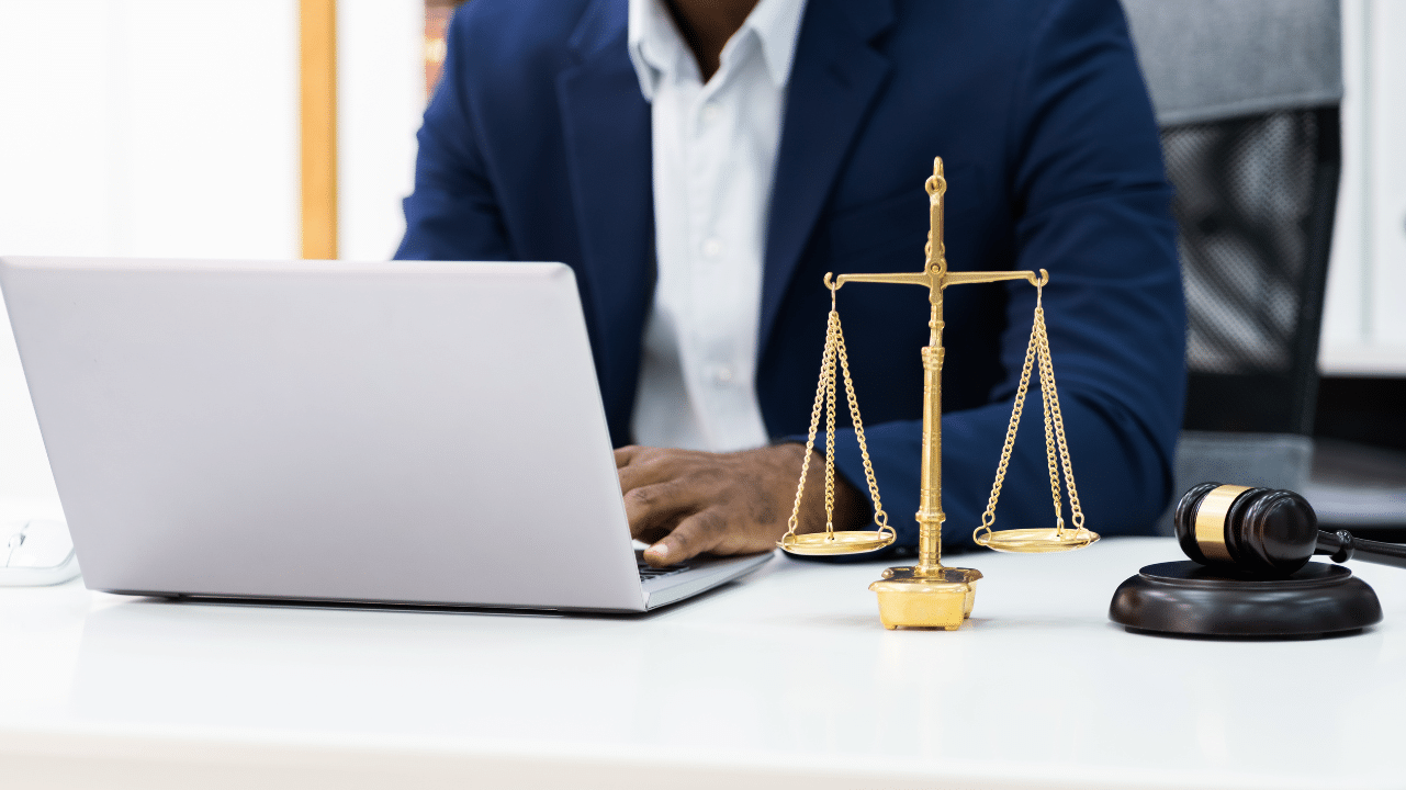 African American man lawyer working on a laptop wearing a dark blue jacket. Justice scale and gavel are next to the laptop.