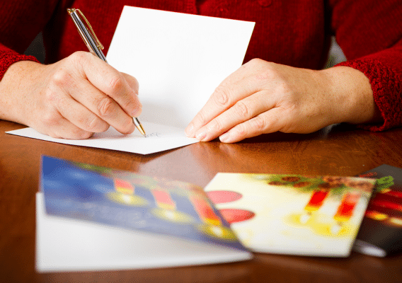 Person in a red knit sweater sitting at a table writing in a card. 3 other cards are sitting on the table in the foreground.