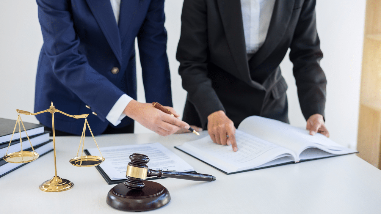 Two professionals dressed in suits pointing at a paper on a desk with a gavel and scales of justice off to the side.