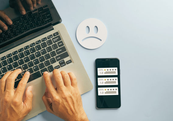 person typing on laptop next to phone displaying 3 one-star reviews and a paper frowning face on the desk